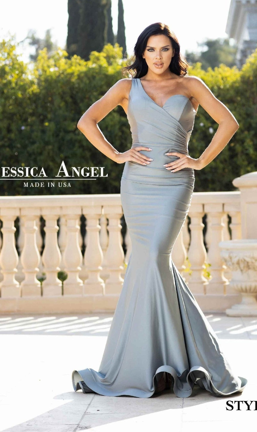 A woman poses outdoors by a stone railing, modeling the elegant Jessica Angel 809—One-Shoulder Long Prom Dress—a stunning, form-fitting mermaid gown in pale blue.